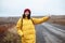 Female tourist with a backpack wearing yellow jacket and red hat catches a car on the road. Young woman travels during winter or