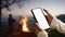 Female tourist hands holding smartphone and rest at night camping beside campfire.