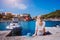 Female tourist sitting on the pier with blau sun hat laying behind. Assos village with beautiful traditional houses is