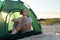 Female tourist sitting in touristic tent with cup of hot tea on sandy beach. Camping