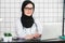 Female veiled scientist sitting on her desk with fingers on a keyboard of her laptop, smiling