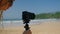 Female vlogger adjusts DSLR on tripod at sunny beach for content creation. Curly hair influencer films tutorial