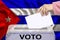 Female voter drops a ballot in a transparent ballot box against the background of the Cuba national flag, vote in spanish, concept