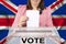 Female voter drops a ballot in a transparent ballot box against the background of the national flag of Great Britain, concept of