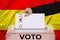 Female voter drops a ballot in a transparent ballot box against the background of the national flag of Spain, concept of state