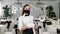 A female waiter in a white shirt and a protective medical mask in the restaurant hall shows a raised thumb