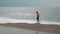 Female is walking on empty sandy beach at stormy windy day, side view tracking shot. Ocean waves are splashing and creating white