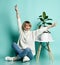 Female in white sweater, jeans, sneakers. Smiling, raised hands, sitting on floor. Chair, green ficus. Posing on blue background