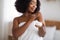 Feminine black woman holding jar of body or face cream, applying skin care product after shower at home