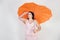 Feminine woman with plus size body in pink dress with orange big heart shaped umbrella posing on white background in Studio