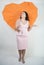 Feminine woman with plus size body in pink dress with orange big heart shaped umbrella posing on white background in Studio