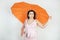 Feminine woman with plus size body in pink dress with orange big heart shaped umbrella posing on white background in Studio