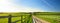 Fence casting shadows on a road leading to small house between scenic Cornish fields under blue sky, Cornwall, England