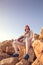 Fencer man sitting on top of the rock holding his fencing mask and a sword on a blue sky background and looking forward