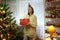 Festive funny girl holding Christmas gift box in living room by decorated living room