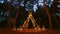 Festive string lights illumination on boho tipi arch decor on outdoor wedding ceremony venue in pine forest at night