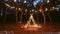 Festive string lights illumination on boho tipi arch decor on outdoor wedding ceremony venue in pine forest at night