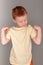 Fiery redhead young boy in yellow shirt looking at left arm with strong arms pose. Portrait on grey with copy space above.