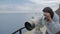 Fifteen-year-old boy in grey hoodie looking at the sea into the telescope from the high elevated observation deck.