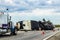 Fifth wheel RV overturned on highway with wench truck trying to get it off the road and two semis parked nearby and traffic cones