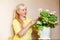 Fifty-year-old beautiful Caucasian woman in a yellow jacket and a blooming pink pelargonium in a pot on the table