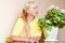 A fifty-year-old beautiful Caucasian woman in a yellow jacket sits at a table next to a pot of geranium blossoms. grow potted