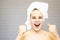 Fighting young woman wrapped in bath towels with cream on face after shower in bath