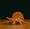 A figurine of an aggressive growling orange dinosaur of the Dimetrodon species stands on a brown table against a dark background