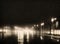 film noir style image of a wet road in the rain at night with vehicle headlights and surrounding buildings reflected on the