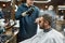 Finishing touch. Barber fixing with hairspray new haircut of young bearded man sitting in barber shop chair. Man