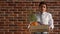 A fired employee with a box of things and a flower inside, stands against a brick wall and shrugs