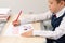 A first grader boy draws a postcard for the holiday of halloween with a red bright pencil on a white sheet of paper while sitting