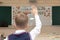 First grader boy at his desk in the classroom on the day of knowledge at school. There is an inscription on the blackboard - for t