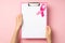 First person top view photo of hands holding pink clipboard with paper sheet and pink ribbon symbol of breast cancer awareness on