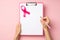 First person top view photo of hands holding pink pen and pink clipboard with paper sheet and pink ribbon symbol of breast cancer