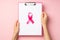 First person top view photo of woman`s hands holding pink clipboard with paper sheet and pink ribbon symbol of breast cancer