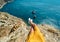 First person view of taned woman legs in yellow dress sitting on on cliff edge with amazing nature view of seascape.