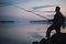 Fisher man fishing with spinning rod on a river bank at misty foggy dusk