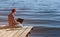 Fisherman man works on a laptop, sits on a wooden pier near the lake, next to it there is a fishing pole.