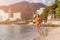 Fit female athlete wearing bikini running on beach with sun shining in camera and hotel resort hills in background