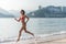 Fit female athlete wearing bikini running on beach with sun shining in camera and hotel resort hills in background