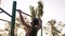 Fit man in black shirt doing pull-ups on horizon bar on sports ground with trees and sun shines on the background