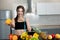 Fit woman with centimeter in one hand stretching out orange wearing black sports top standing in the kitchen full of fruits,