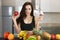 Fit young woman with apple in one hand and cream cake in another standing in kitchen full of fruits looking hesitant dietology and