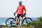 Fitness man with a bicycle stands on the ocean shore and looks into the distance on a sunny day.