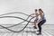 Fitness people exercising with battle ropes at gym. Woman and man couple training together doing battling rope workout