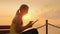 Fitness woman enjoys a tablet. Sits on a bench on a pier against the backdrop of the rising sun