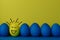 Five blue and one yellow funny faced painted Easter eggs stand in a row on a yellow with blue background.