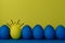 Five blue and a yellow funny faced painted Easter eggs stand on a yellow with blue background. Creative Easter concept