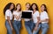 Five multiethnic women holding laptop over yellow studio background, mockup